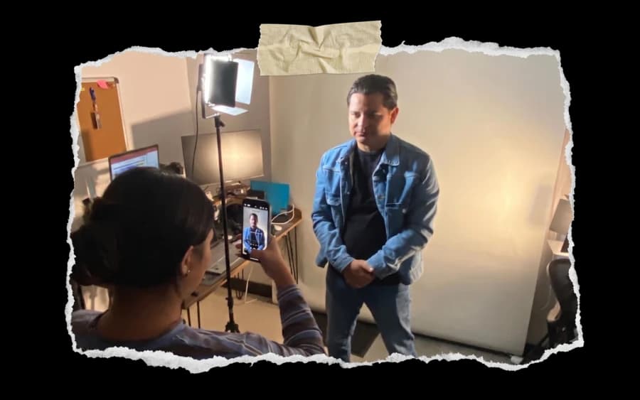Sesión de retrato en estudio: una integrante del equipo fotografía con el celular a un entrevistado de pie frente a un fondo blanco iluminado.